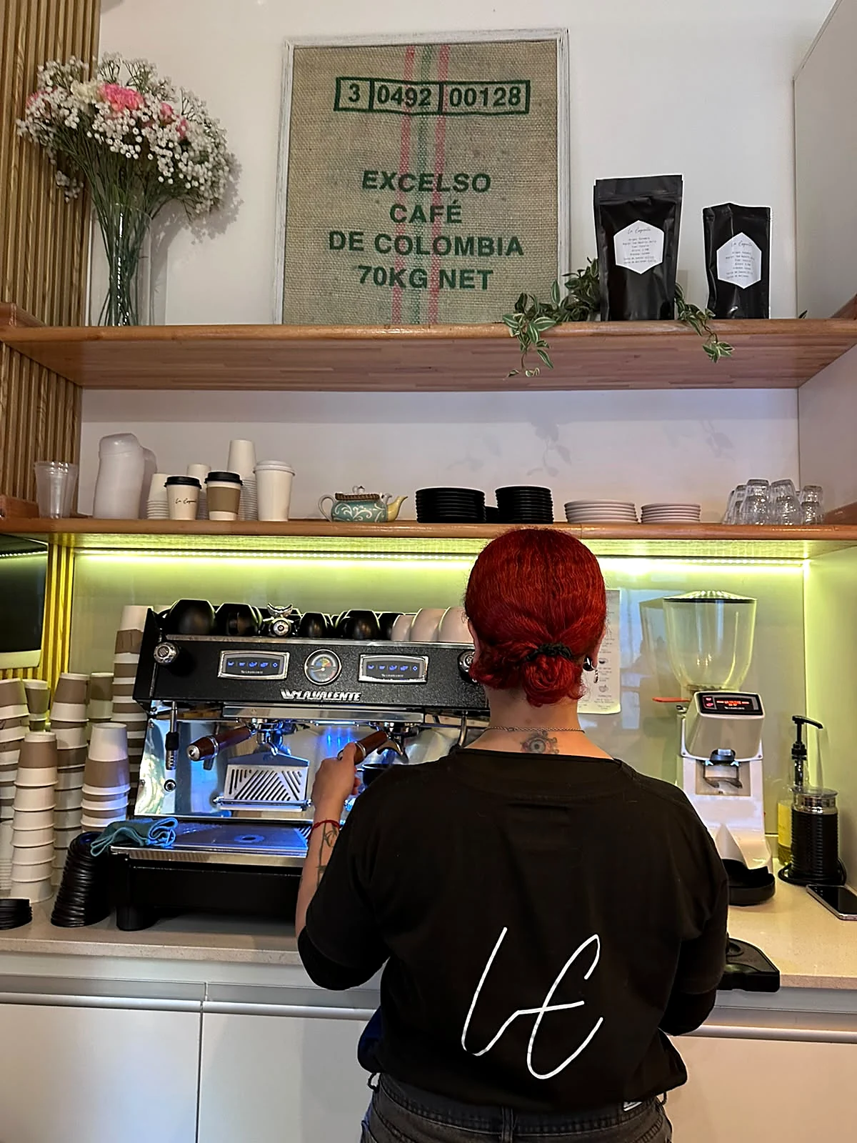 Barista de espaldas con el LE en la remera, preparando café en la máquina de espresso; arriba, un saco de café Excelso de Colombia